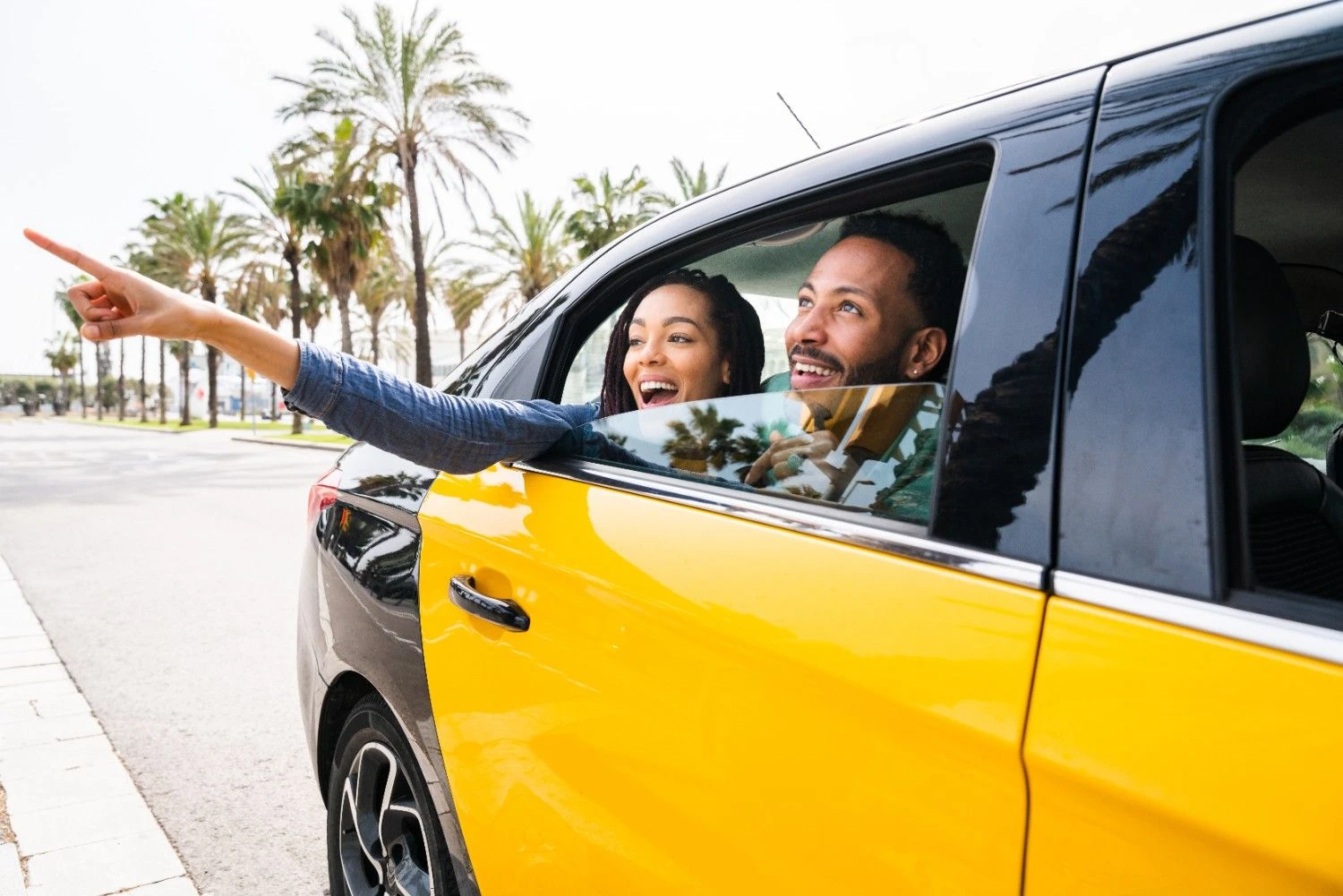 Two people leaning out of a yellow car window and pointing ahead on a palm lined road
